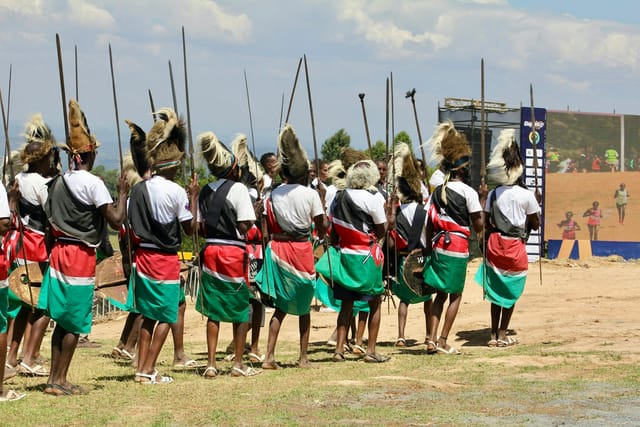 Warriors in traditional attire holding long spears, with shields among the group.